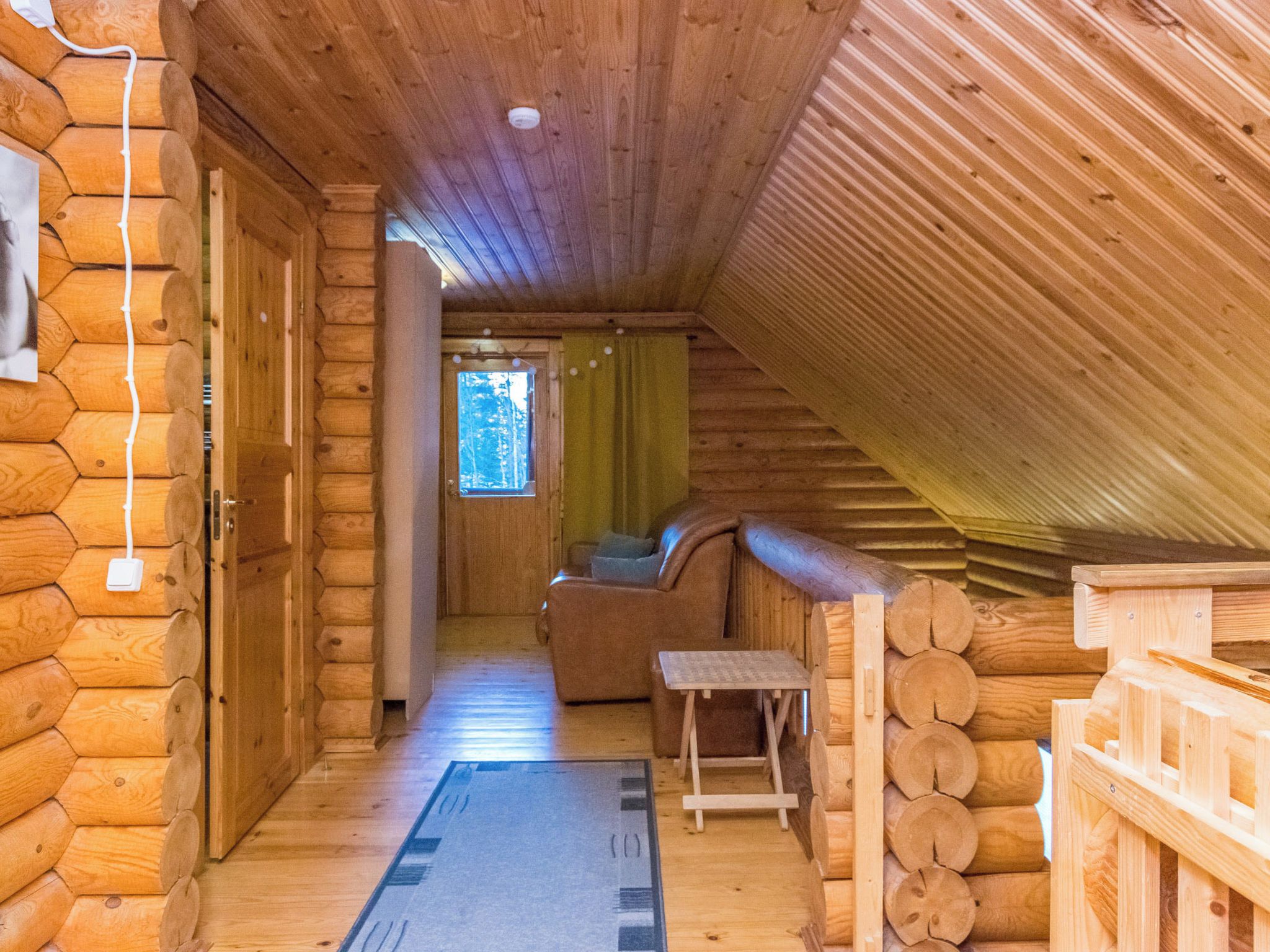 A log cabin interior with a wooden staircase, a sofa, a small table, and a window with a view of trees.