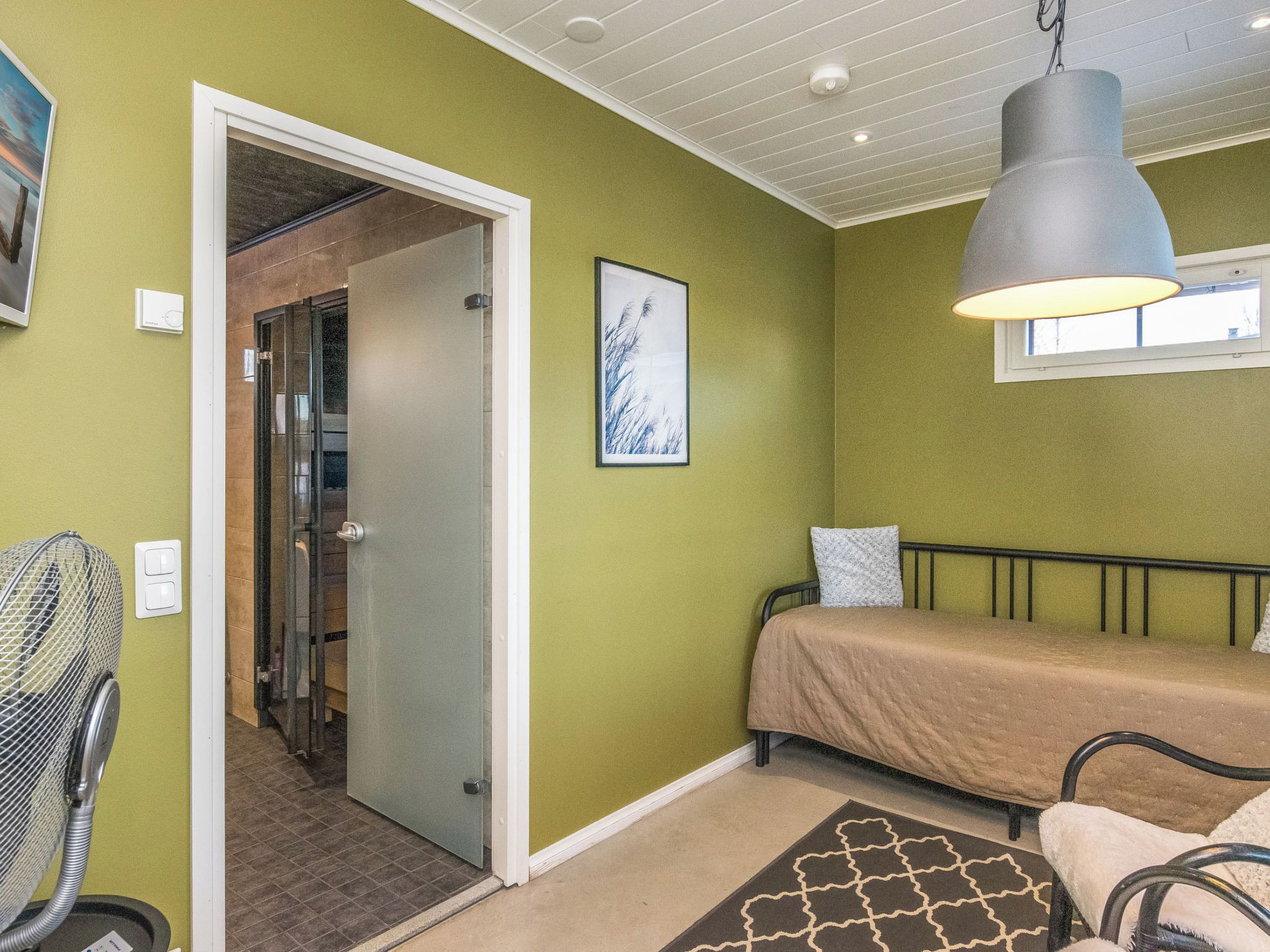 A room with a bed, a chair, a standing fan, a picture on the wall, and a door leading to another room.