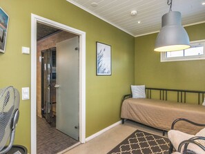 Une pièce comprenant un lit, une chaise, un ventilateur sur pied, un tableau accroché au mur et une porte donnant sur une autre pièce.