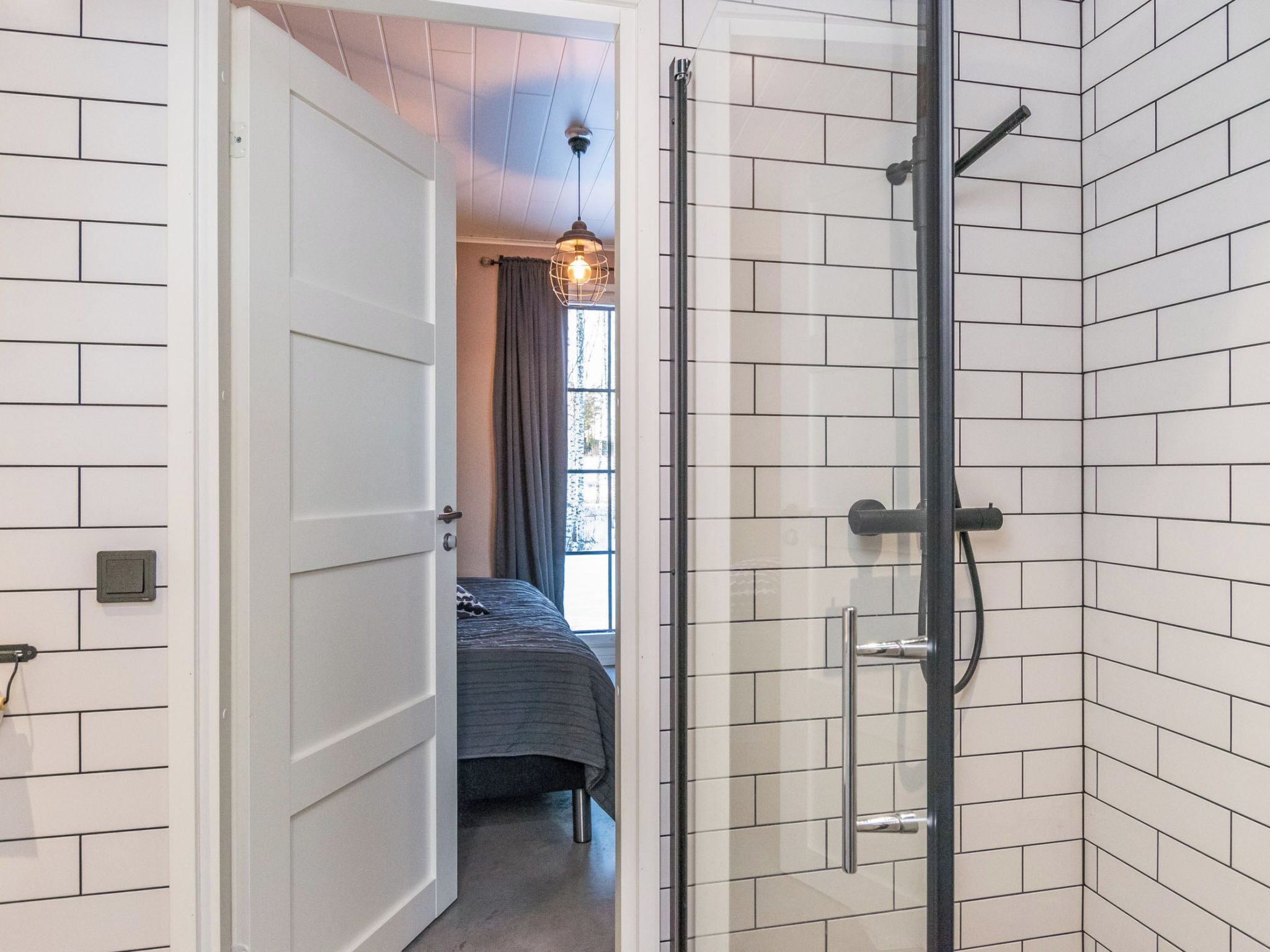 Ein Badezimmer mit weiß gefliesten Wänden, einer Duschkabine aus Glas und einem sichtbaren Bett im angrenzenden Raum.