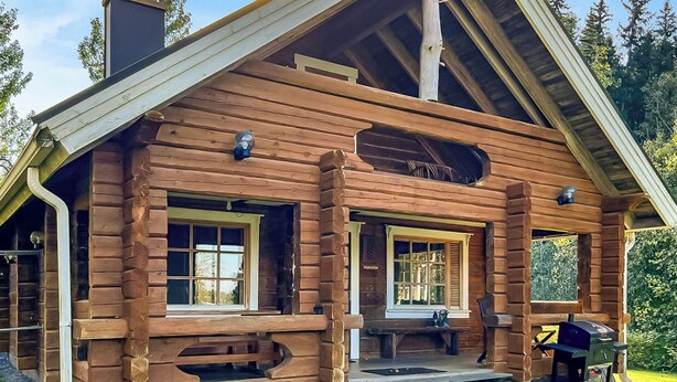 Een vakantiehuis in blokhutstijl met een steile puntige dakschild, grote ramen en een veranda met een barbecue.