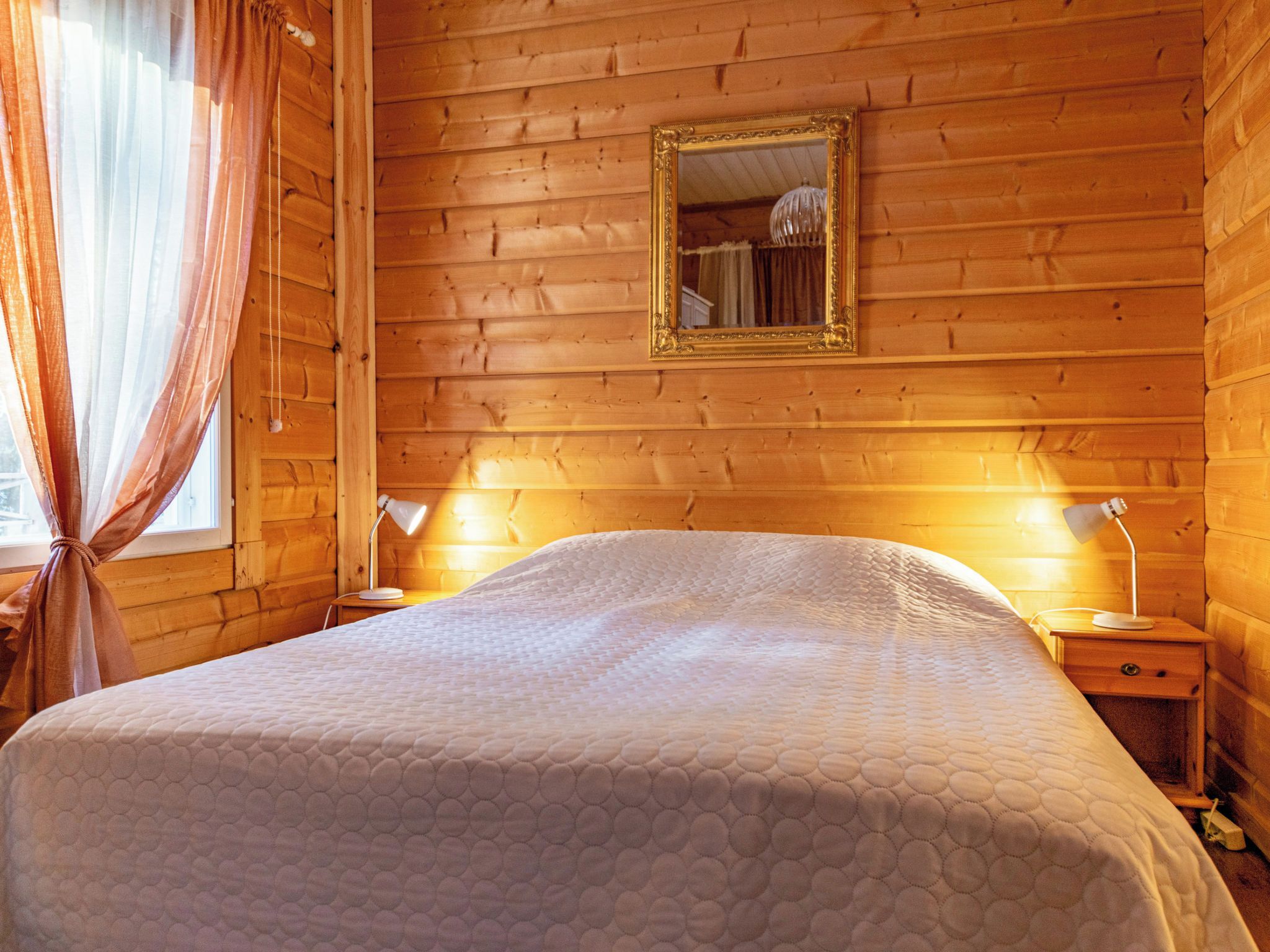 A wooden-paneled room with a bed, two bedside lamps, a mirror, and a window with curtains.