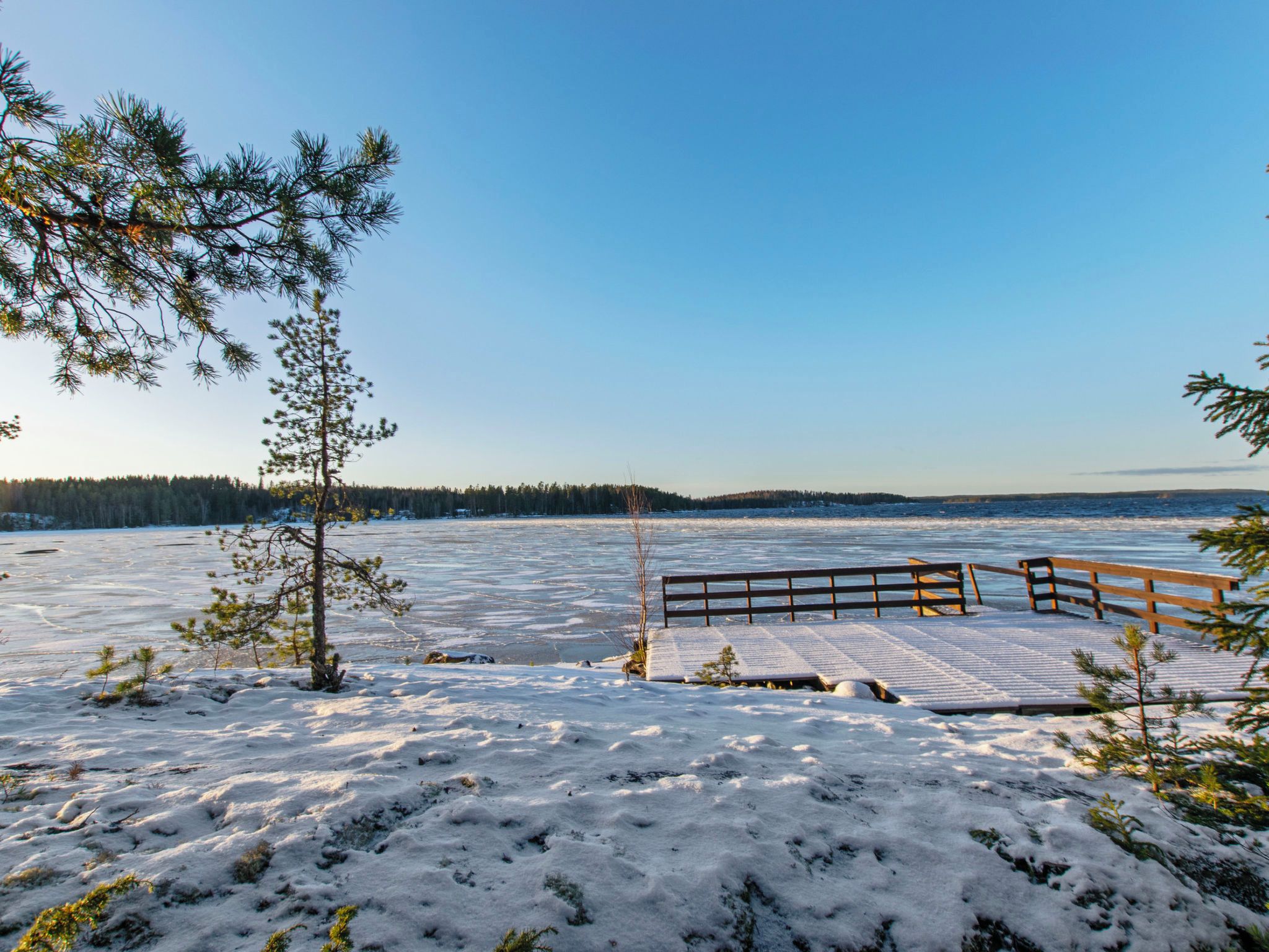A snowy landscape with a wooden dock extending into a frozen lake, surrounded by sparse trees.