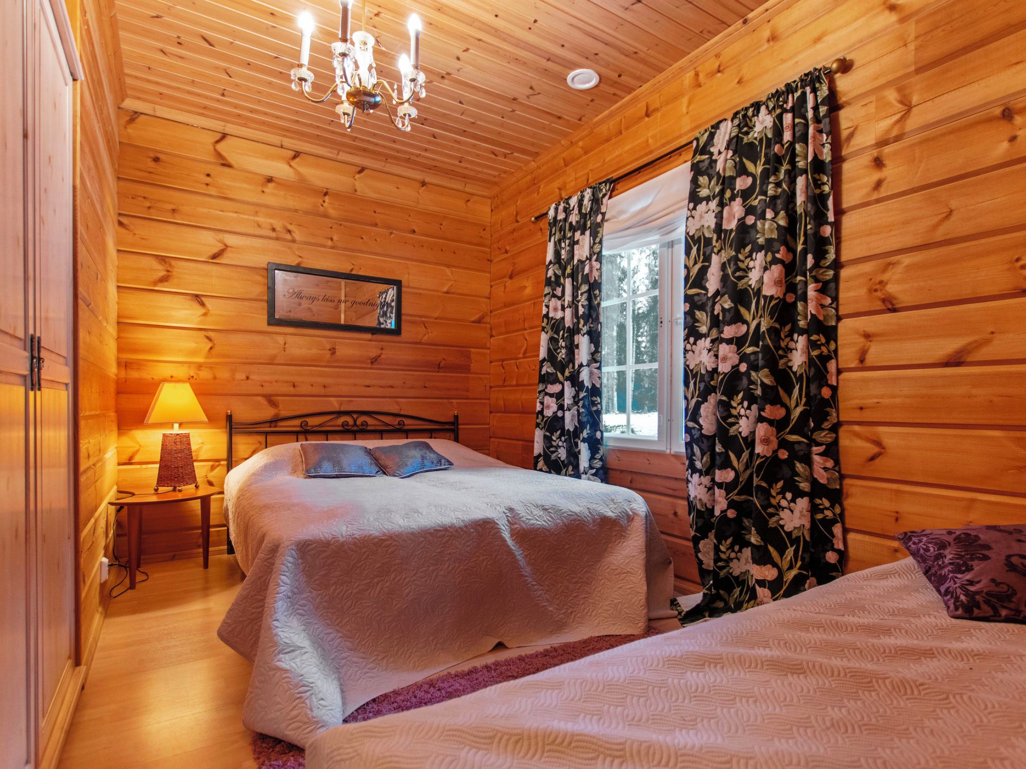 A wooden cabin bedroom with two beds, a chandelier, a bedside table with a lamp, and a window with floral curtains.