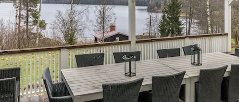 An outdoor deck with a table and chairs, overlooking a lake with trees and a cabin in the background.