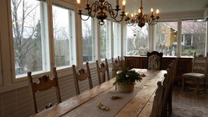 A dining room with a long wooden table, multiple chairs, a chandelier, and large windows with a view of trees and a house.