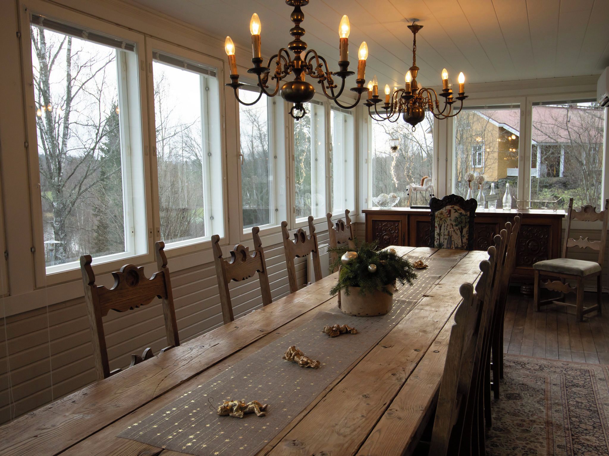 A dining area with a long wooden table, chairs, a chandelier, and large windows.