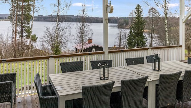 A covered outdoor deck with a table and chairs, overlooking a lake and forest.