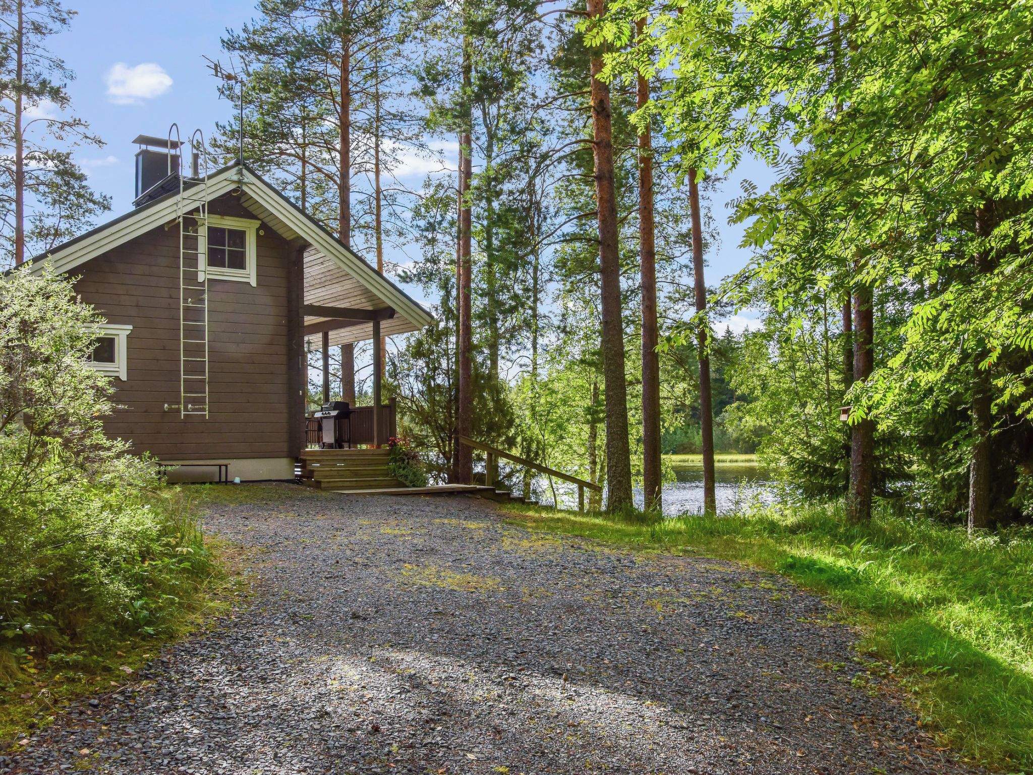 Mökki, jonka edessä on sorapäällysteinen ajotie, joka johtaa puiselle terassille. Mökkiä ympäröivät korkeat puut ja sen lähellä on järvi.