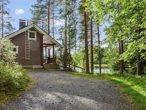 A cabin with a gravel driveway leading to a wooden deck, surrounded by tall trees and a lake.