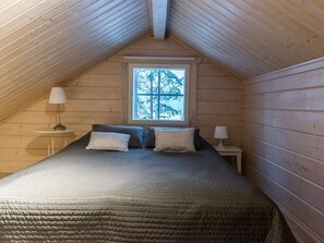 Une chambre confortable située sous les toits, avec un plafond en pente, une grande fenêtre donnant sur les arbres, un lit agrémenté d’oreillers à rayures et deux lampes de chevet.