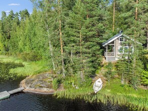 Een chalet omgeven door dicht bos, een steiger die uitkomt bij een vijver, en een klein bootje.