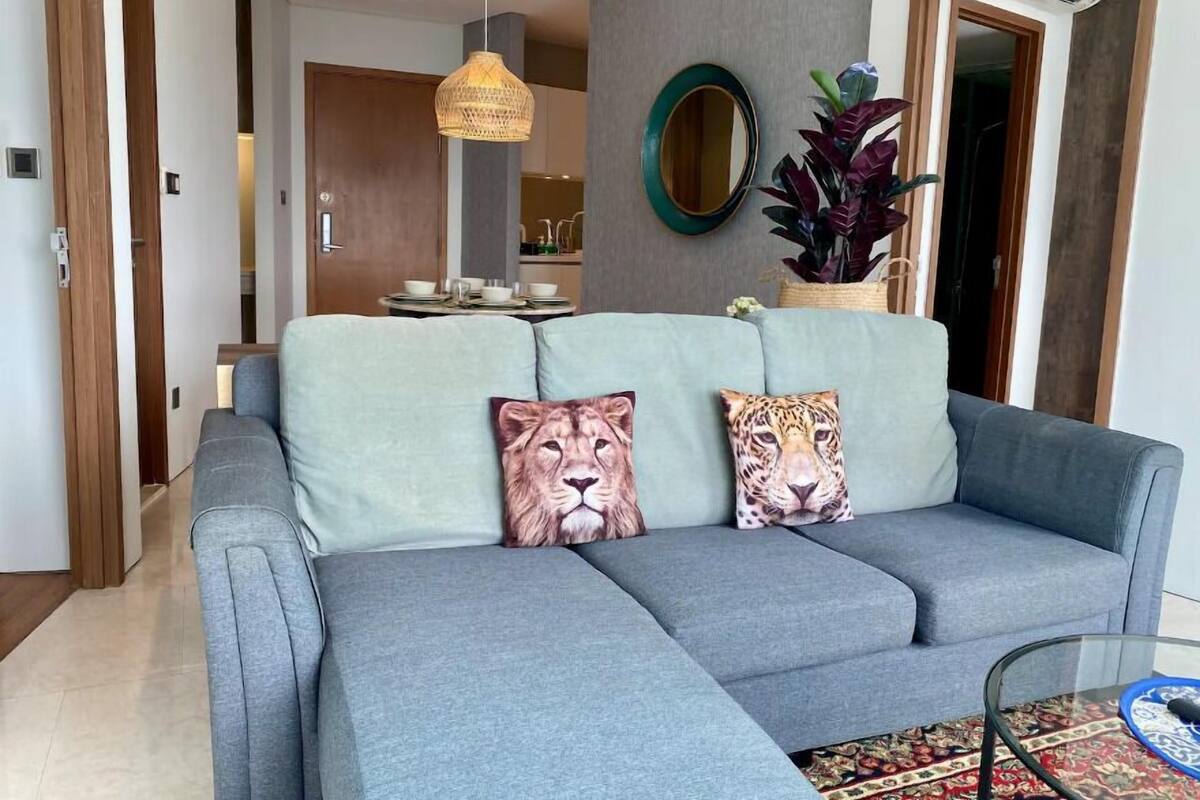 A modern living room with a grey sectional sofa, two leopard-themed cushions, a woven pendant light, a round mirror, and a potted plant.