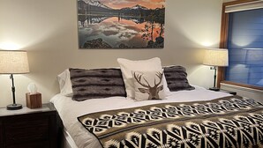 A neatly arranged bedroom with a large bed, two bedside tables with lamps, a wall-mounted painting, and a window with blinds. - Recently Remodeled Golf Course Condo w/ A/C, SHARC Passes, Bikes & Pet Friendly (Sunriver)