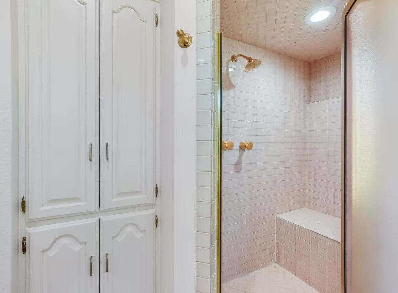 Baño con paredes de azulejos blancos, ducha con cabezal de ducha manual y puerta blanca con pomos de latón.