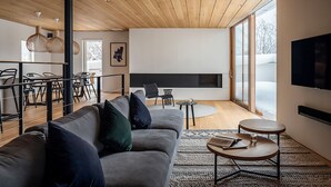 A modern living room with a grey sofa, black and green cushions, a wooden ceiling, and a large window.