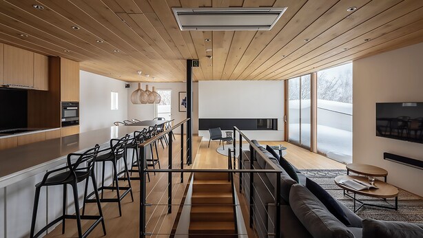 A modern kitchen with wooden cabinets, a central island with bar stools, a dining area with a sofa and coffee table, and a staircase leading to a loft space.