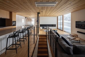 Eine moderne Küche mit Holzausbau, einer zentralen Kochinsel mit Barhocker, einem Essbereich mit Sofa und Couchtisch sowie einer Treppe zu einem Schlafbereich.