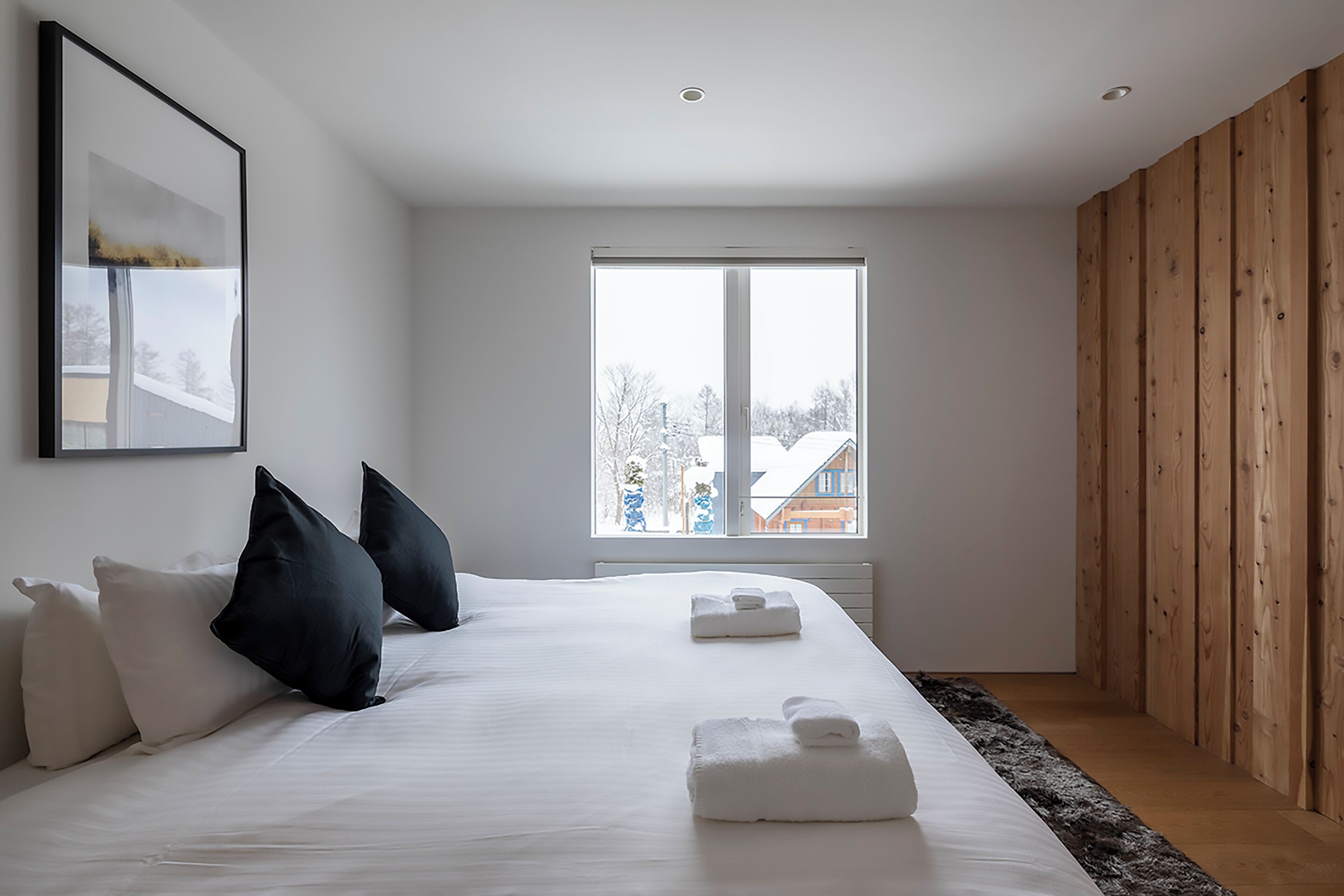 A modern hotel room with a large bed, white linens, a wooden panel wall, and a window with a view of buildings.