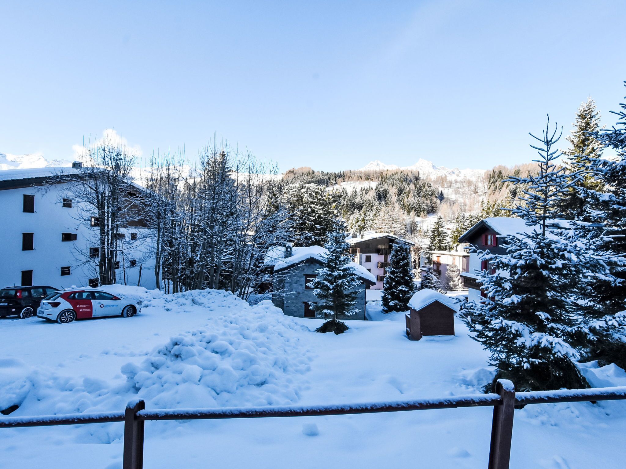 A snow-covered residential area with buildings, a parked car, and a wooden fence.