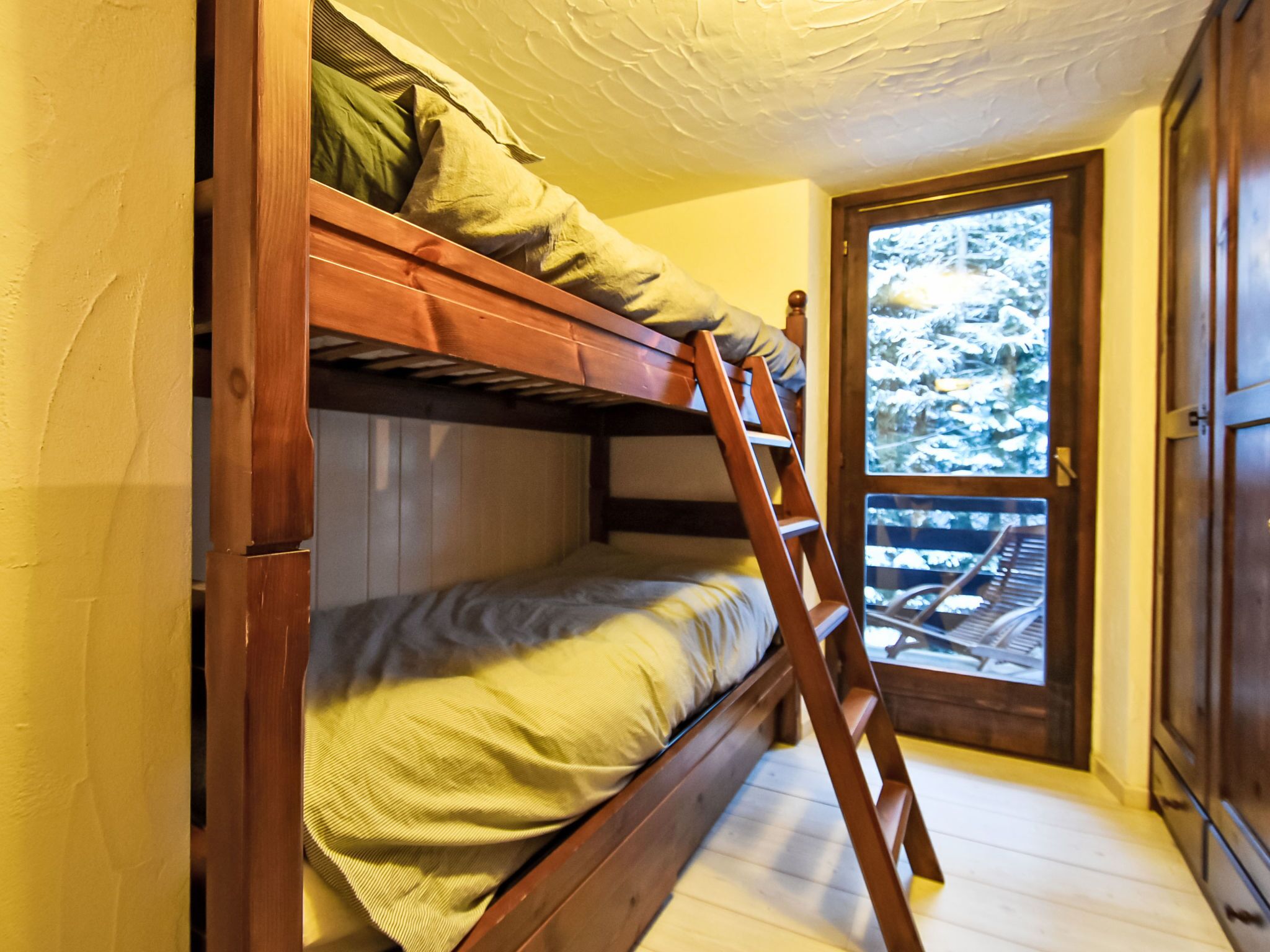 A bunk bed with a wooden ladder, a window with a view of trees, and a wooden door.