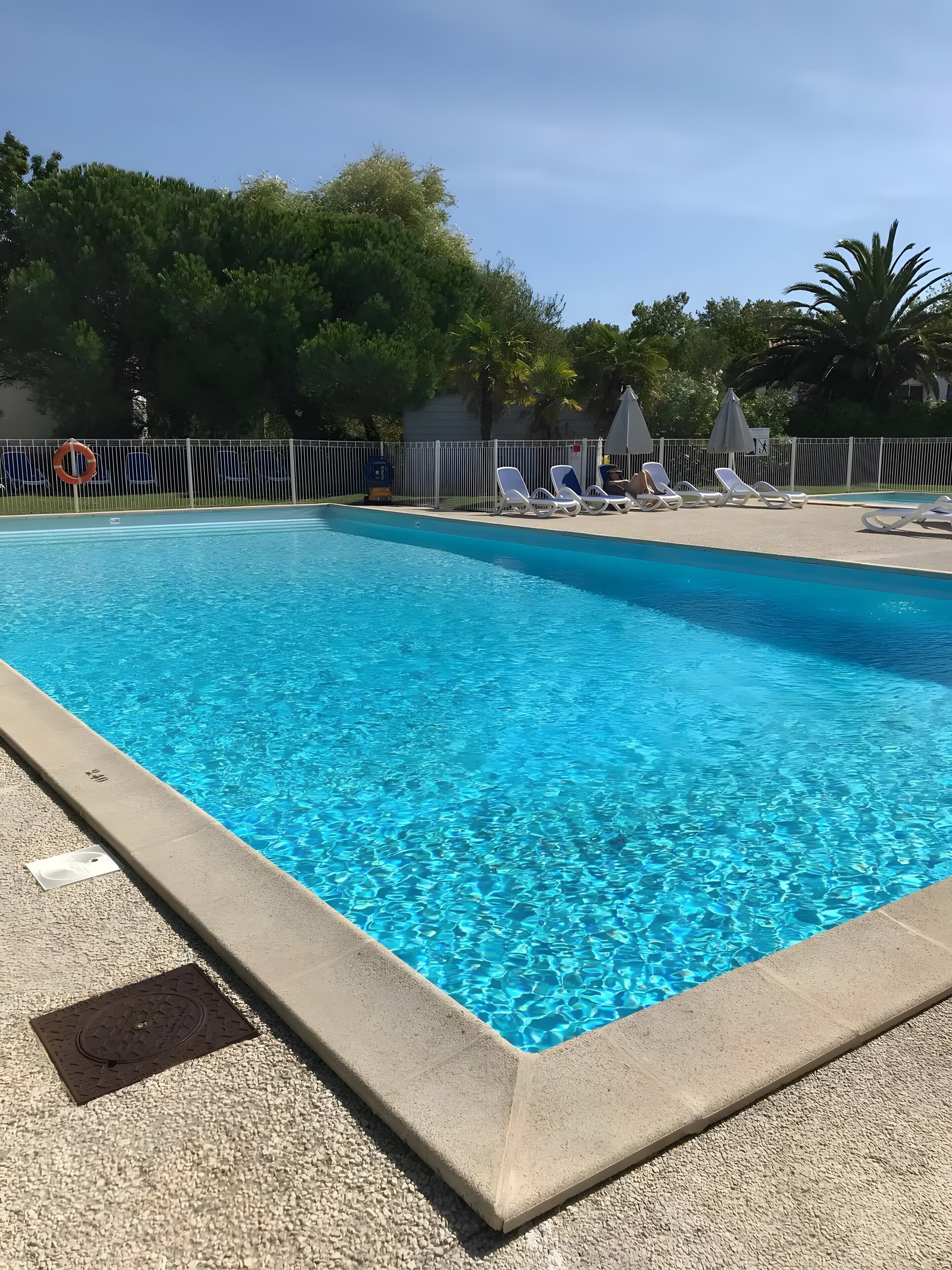 A clear blue swimming pool with lounge chairs and umbrellas, surrounded by a fence and trees.