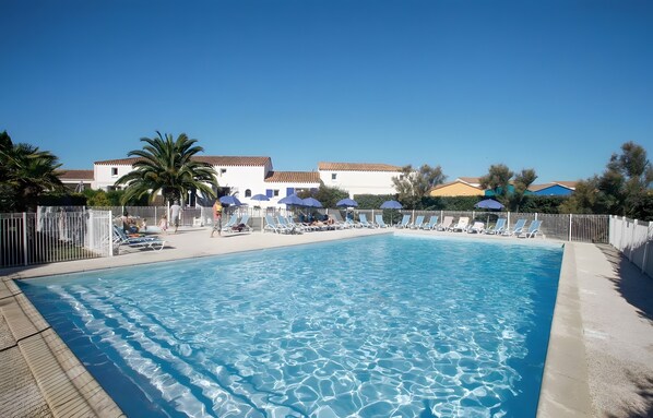 A rectangular swimming pool with clear blue water, surrounded by white sun loungers and umbrellas, with buildings in the background.