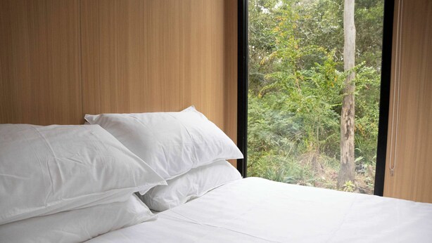 Un lit avec du linge de lit blanc et une vue sur une forêt par la fenêtre.