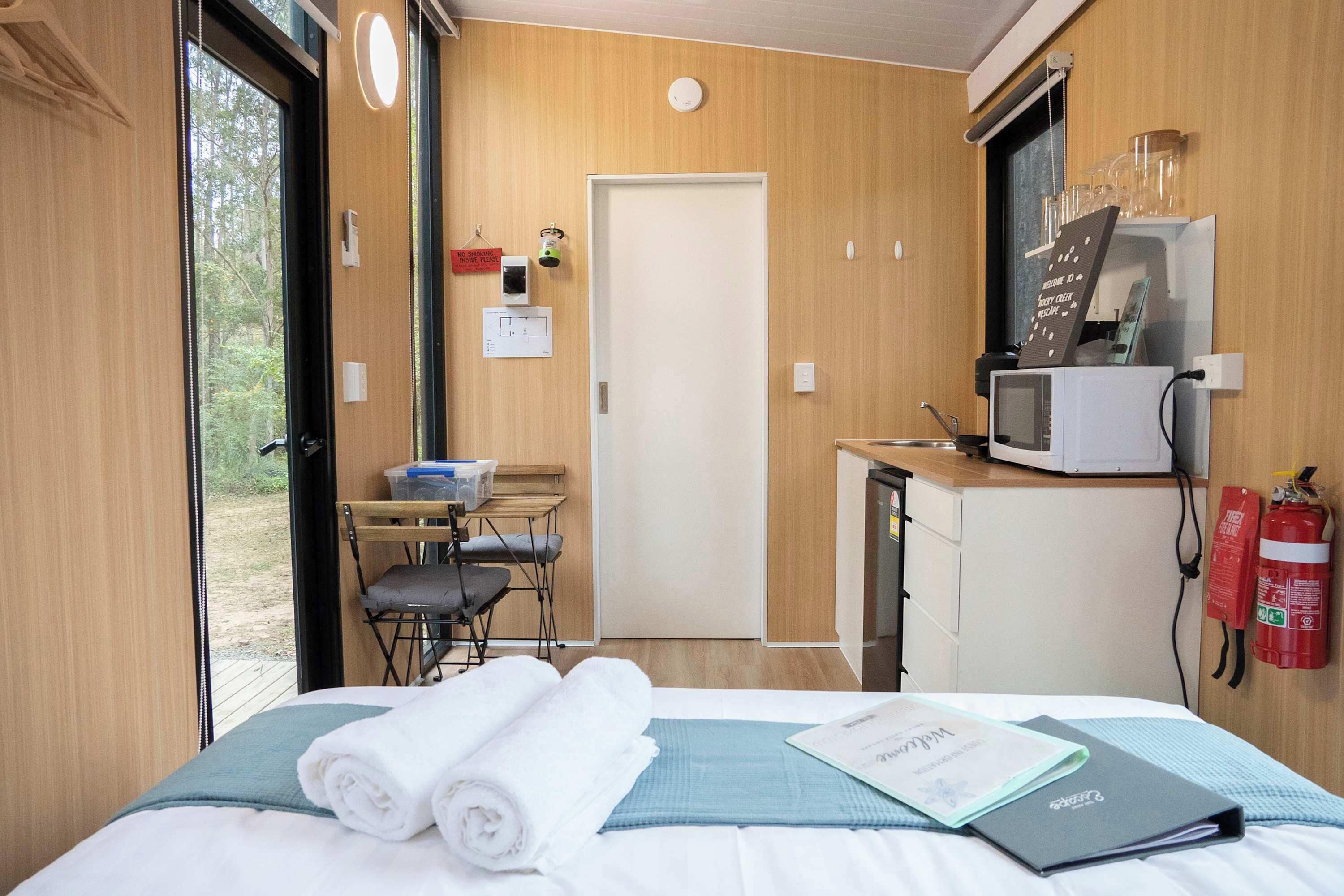 Een compacte kamer met een bed, een klein bureau, een magnetron en een brandblusser.