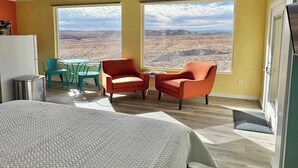 A room with a bed, two chairs, a table, a refrigerator, and large windows with a desert view. - Fossil Knob Ridge -Sierra Vista: Ultra Modern (Terlingua)