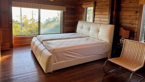 A wooden cabin bedroom with a large bed, two windows, a chair, and a hanging light fixture.