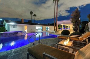 A swimming pool with a fire pit and lounge chairs, surrounded by palm trees and a modern house.