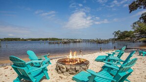A beach with turquoise Adirondack chairs arranged around a stone fire pit, overlooking a calm lake with docks in the distance. - Majestic Retreat - Navarre Townhouse With Bay Access, Garage Community Pool, Kayak, and Paddleboard (Navarre)