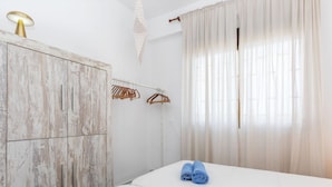 A bedroom with a bed, a wooden wardrobe, a hanging light fixture, and a window with sheer curtains.