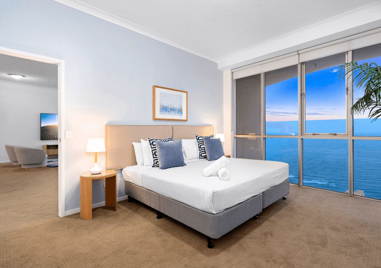 Une chambre d’hôtel avec un grand lit, une table de chevet, une lampe, un tableau encadré au mur et une vue sur l’océan par de grandes fenêtres.