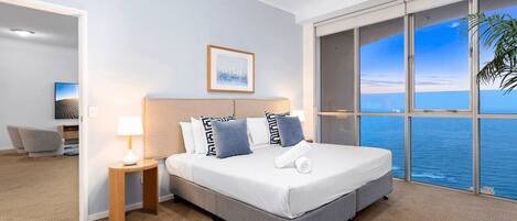 Une chambre d’hôtel avec un grand lit, une table de chevet, une lampe, un tableau encadré au mur et une vue sur l’océan par de grandes fenêtres.