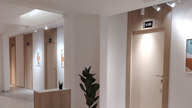 A modern hotel lobby with a reception desk, potted plant, and doors labeled with room numbers. - Private room 8m for men. Bed, AC, side table, Wi-Fi. Shared baths, lounges. (Buraydah)