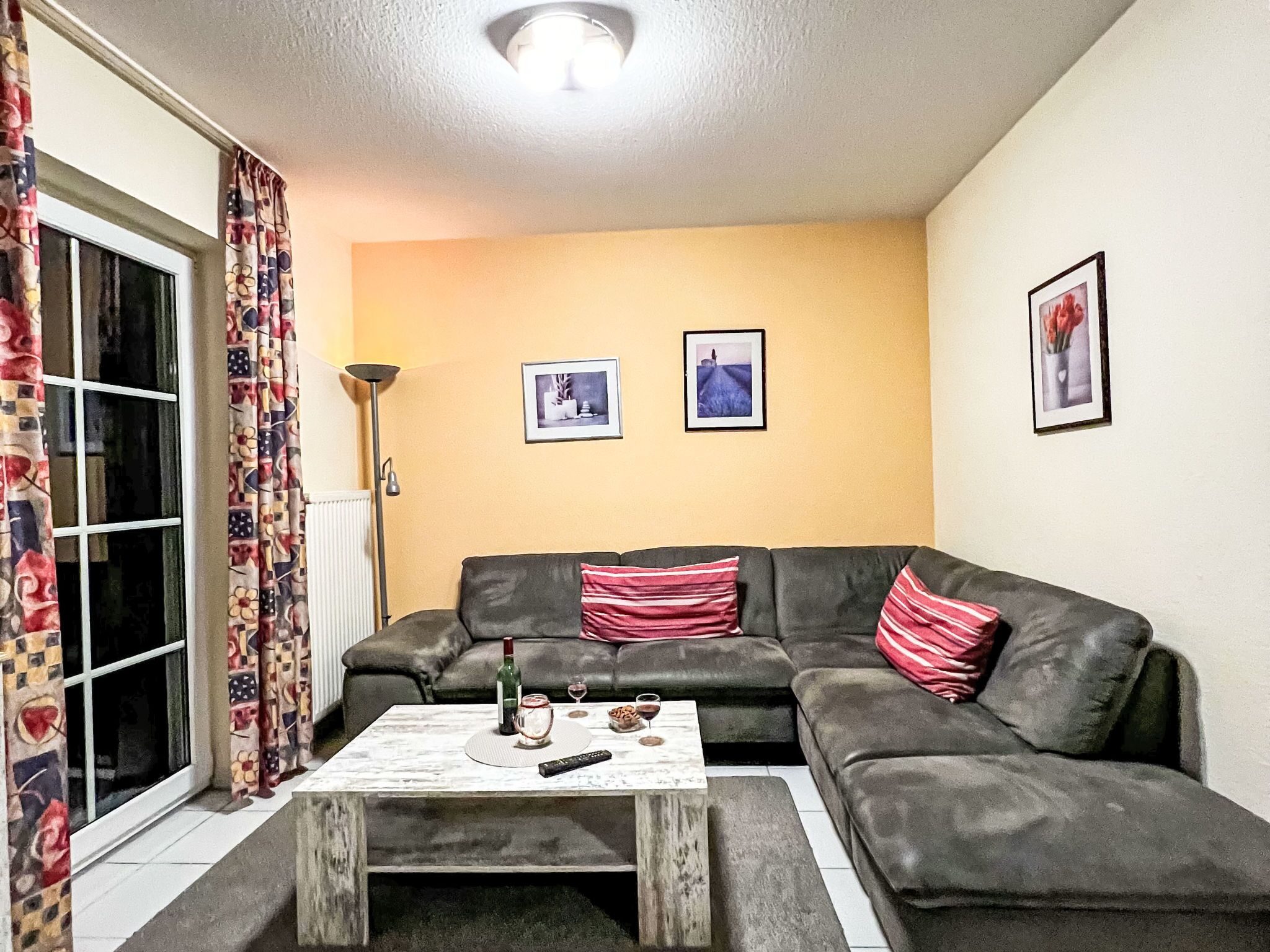 Een woonkamer met een grijze bank, twee rode en witte streepkussens, een salontafel met een fles en glazen, en twee ingelijste foto’s aan de muur.