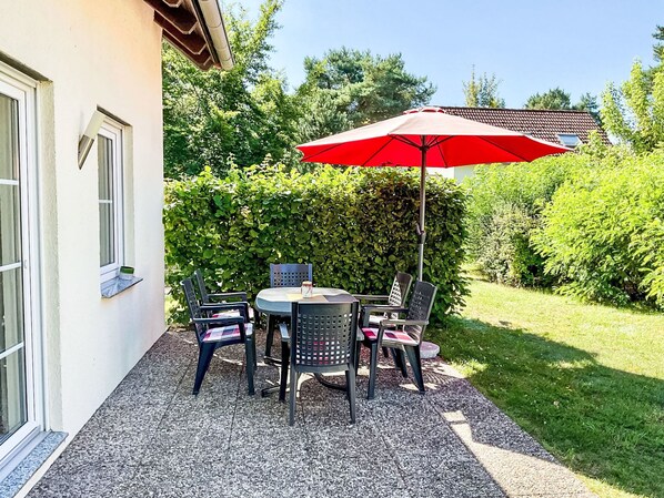 Un patio al aire libre con una mesa, sillas, un paraguas rojo y un seto al fondo.