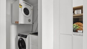 A compact laundry room with a top-loading washer and a front-loading dryer, a wooden shelf with a kettle, and a potted plant.