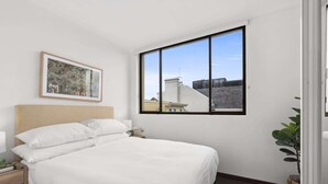 A bedroom with a large window, a bed with white bedding, a bedside table, and a framed picture on the wall.