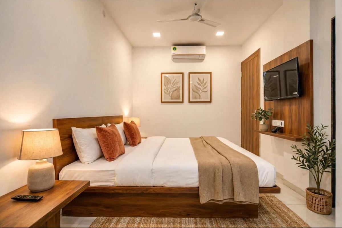 Habitación de hotel moderna con cama, mesita de noche, lámpara, aire acondicionado, televisor montado en la pared y una obra de arte enmarcada.