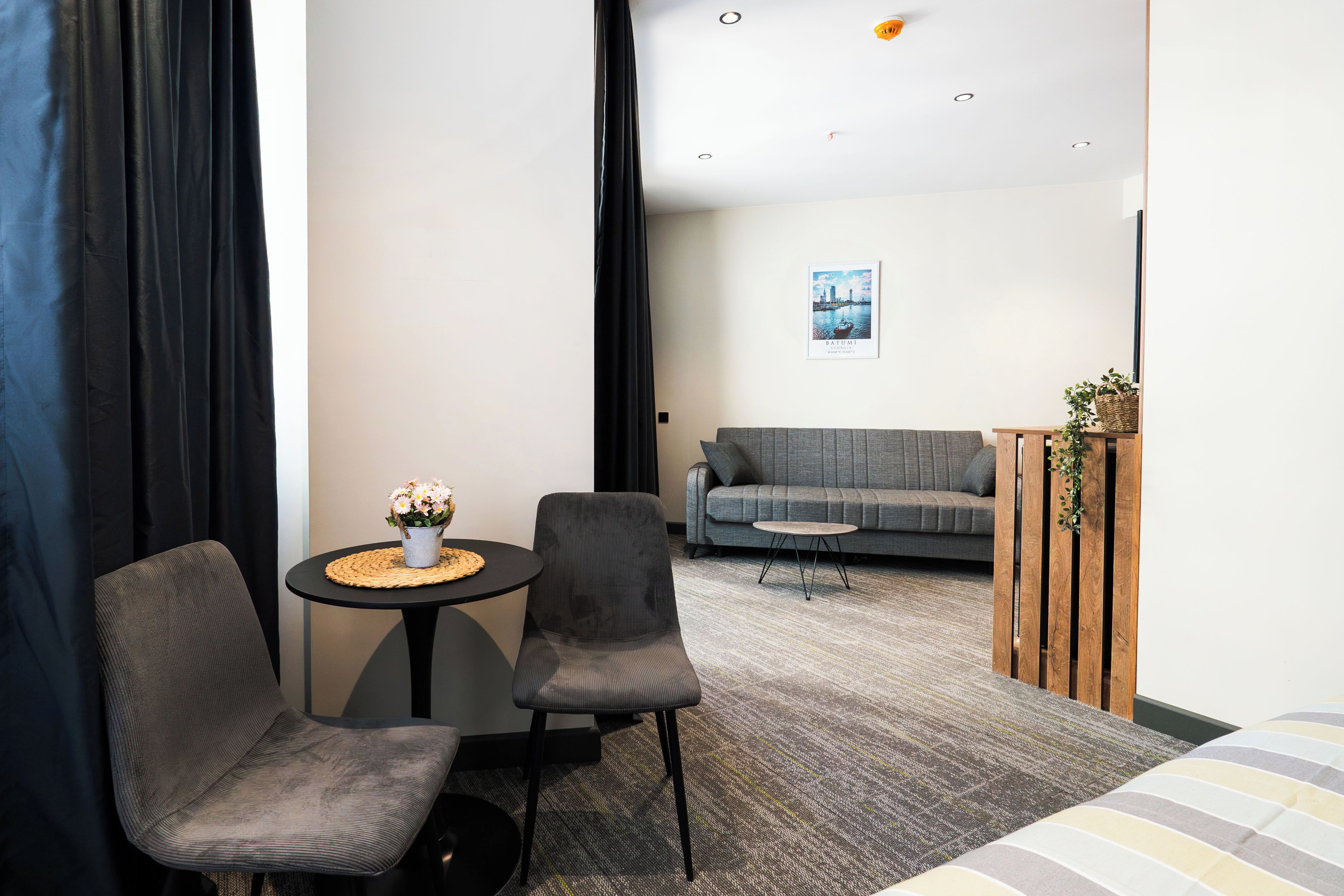 Quarto de hotel moderno com sofá cinza, duas poltronas cinzas, uma mesa redonda pequena com um vaso de flores e uma cama com roupa de cama listrada.