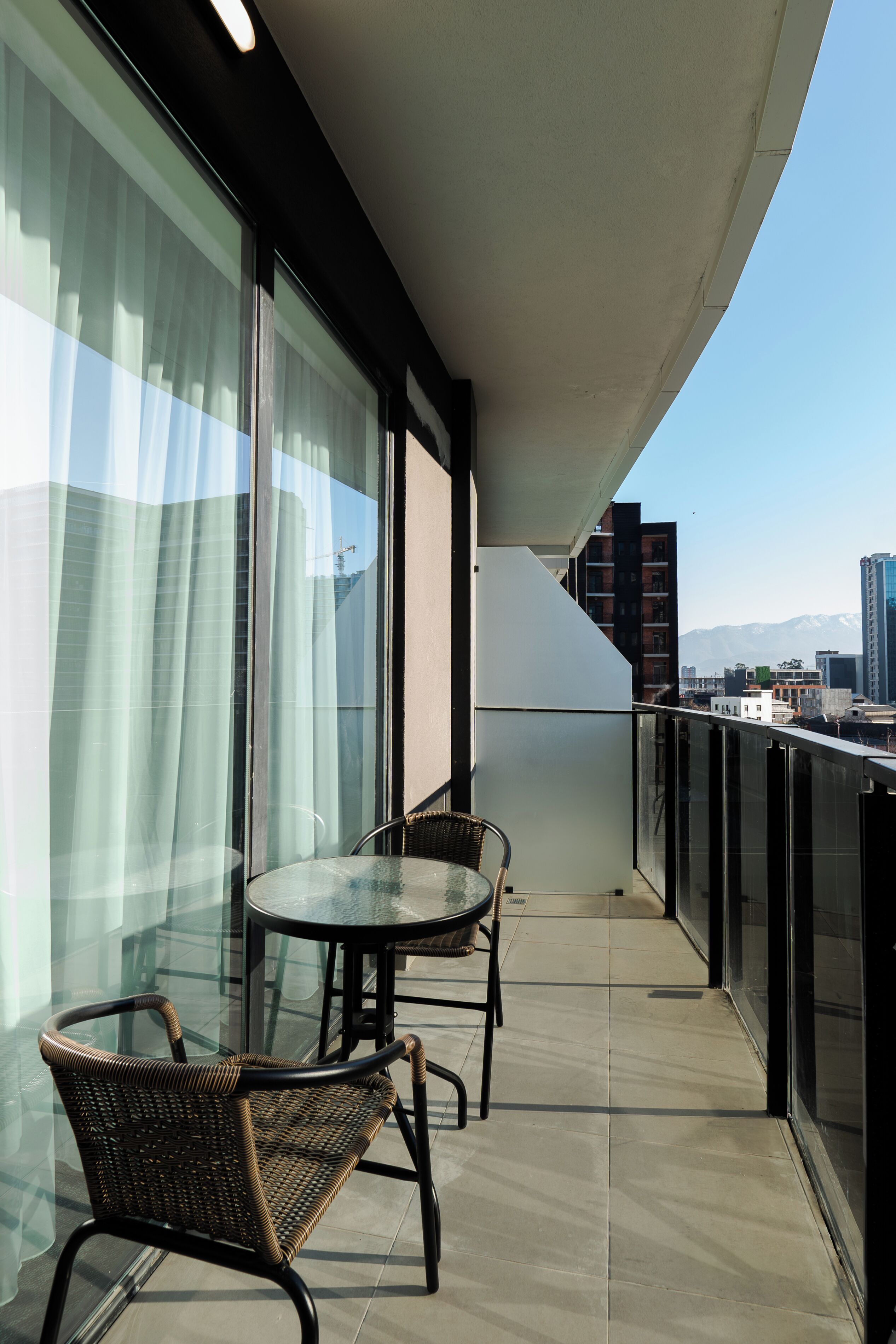 A balcony with two wicker chairs and a small table, large windows with sheer curtains, and a view of city buildings and mountains in the background.