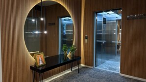 Un área de recepción moderna con un muro revestido de paneles de madera, un ascensor de vidrio y un mostrador de recepción con un cuadro enmarcado.