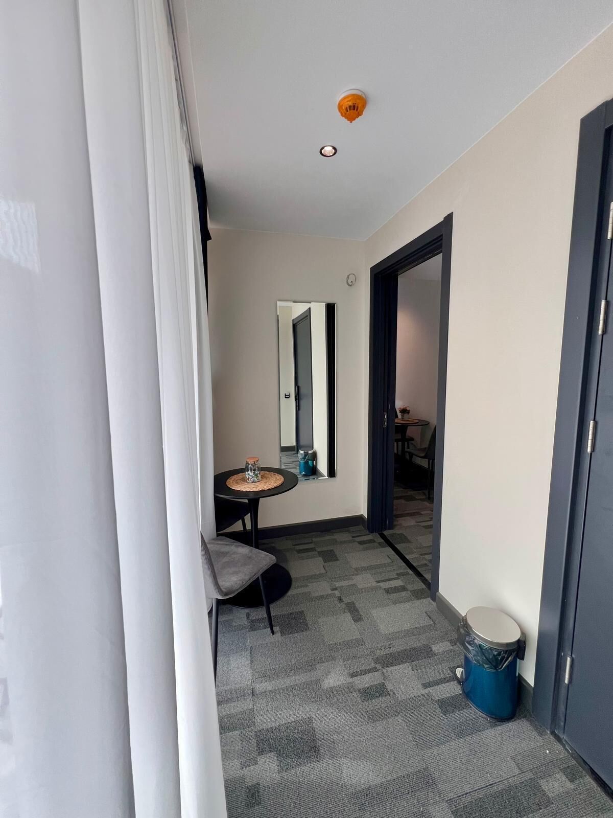A narrow hallway with a small table and chair, a trash bin, and a door leading to another room.