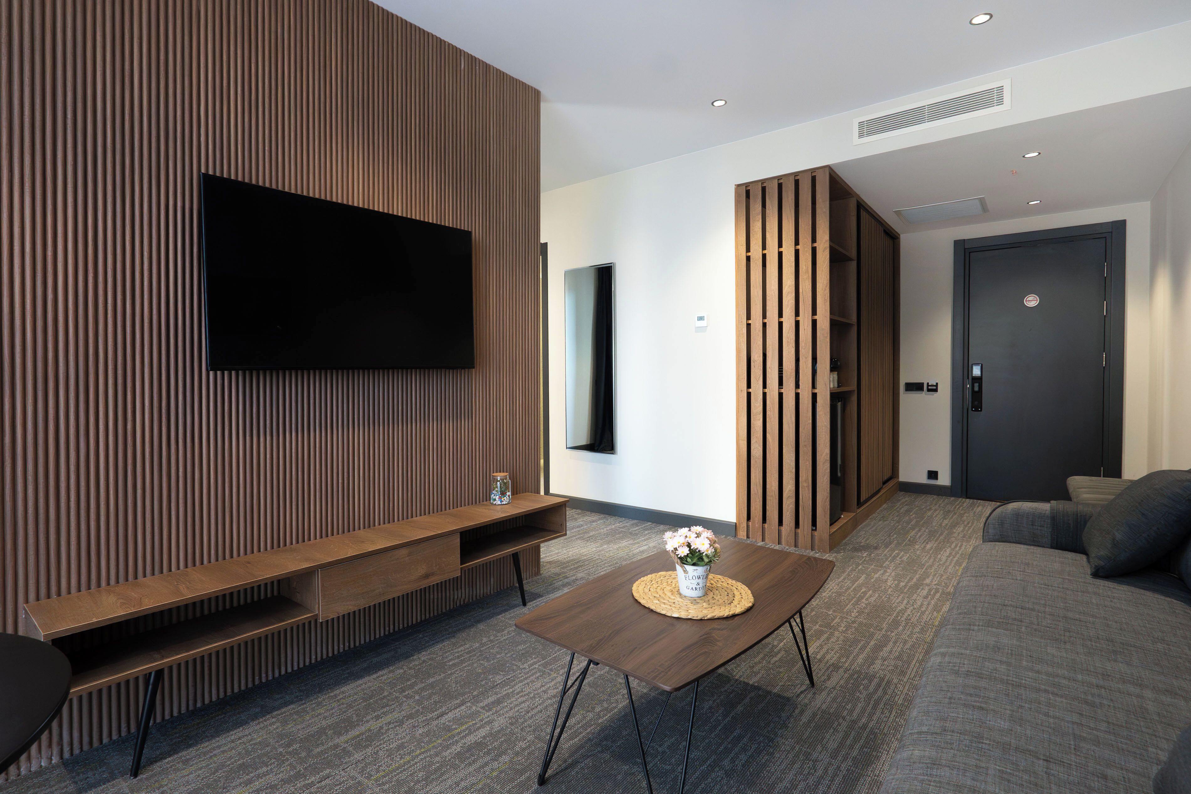 Una sala de estar moderna con un televisor de pantalla plana montado en una pared de madera, un sofá, una mesa de centro con un jarrón y una puerta con cerradura electrónica.