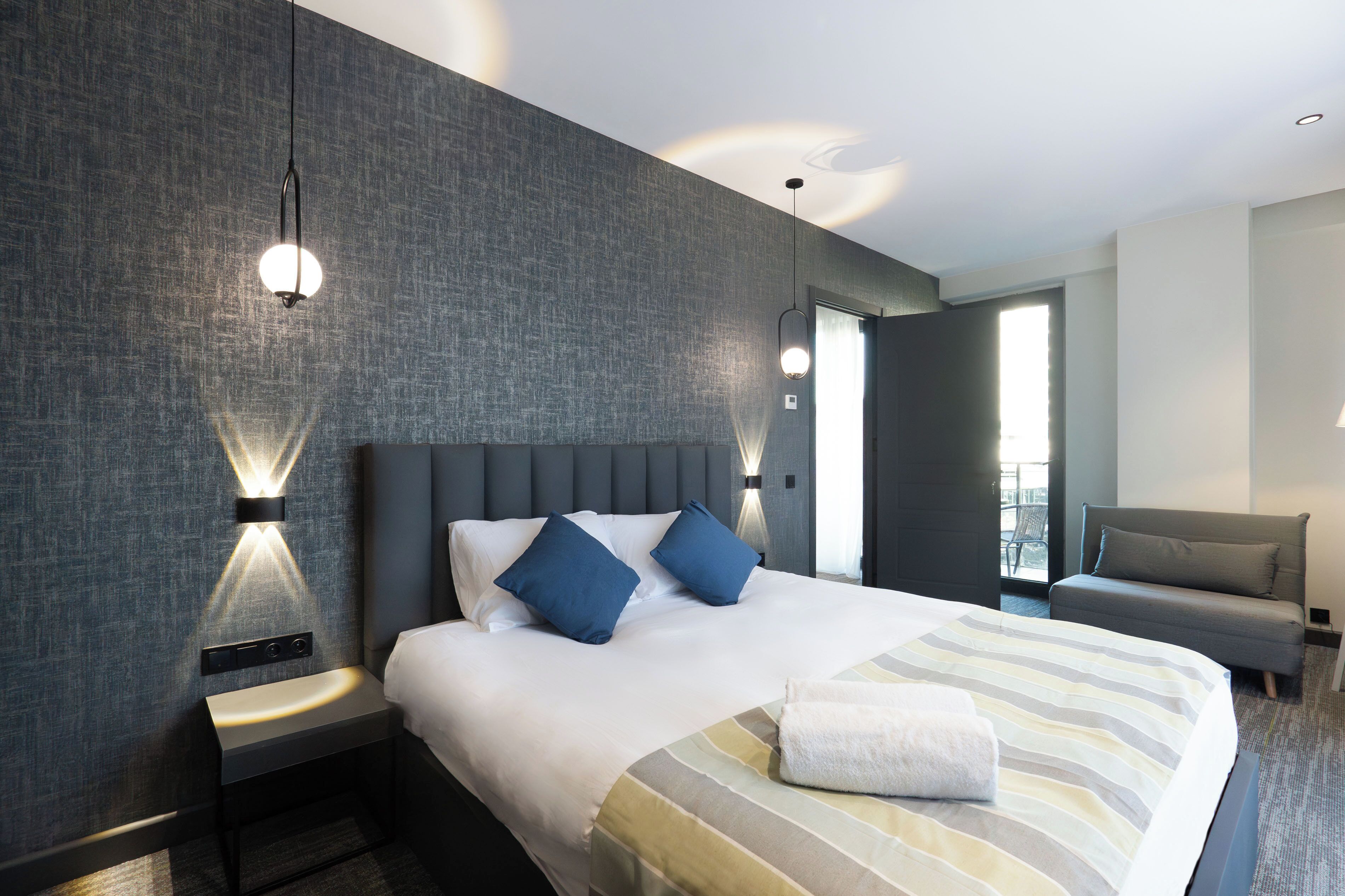 A modern hotel room with a large bed, two bedside tables, a sofa, and a view of the outdoors.