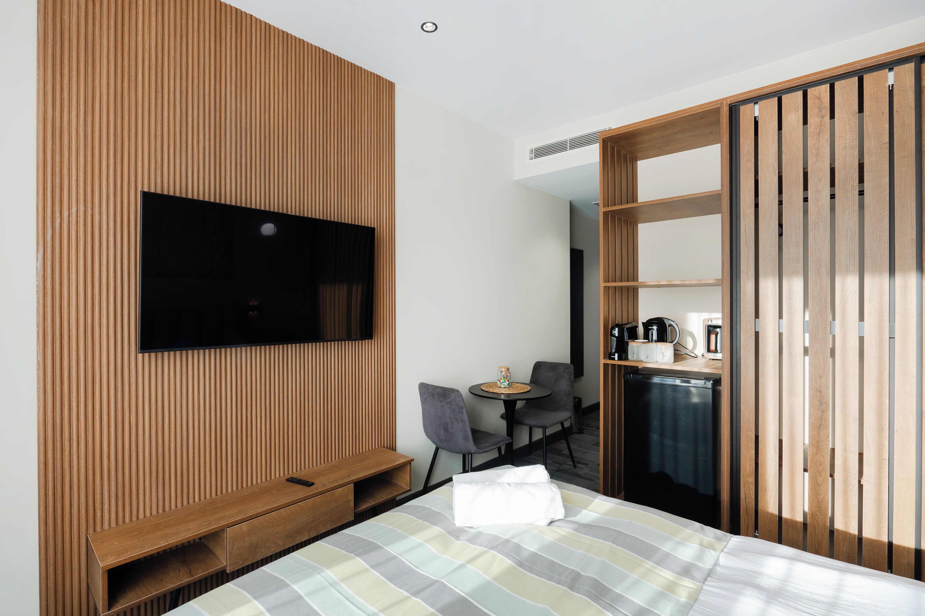A modern hotel room with a large bed, a flat-screen TV mounted on a wooden panel wall, a small round table with two chairs, and a wardrobe with sliding doors.