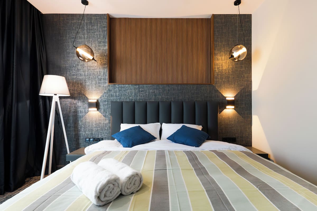 Une chambre d’hôtel avec un lit, deux lampes fixées au mur, une lampe sur pied et une grande fenêtre avec des rideaux.